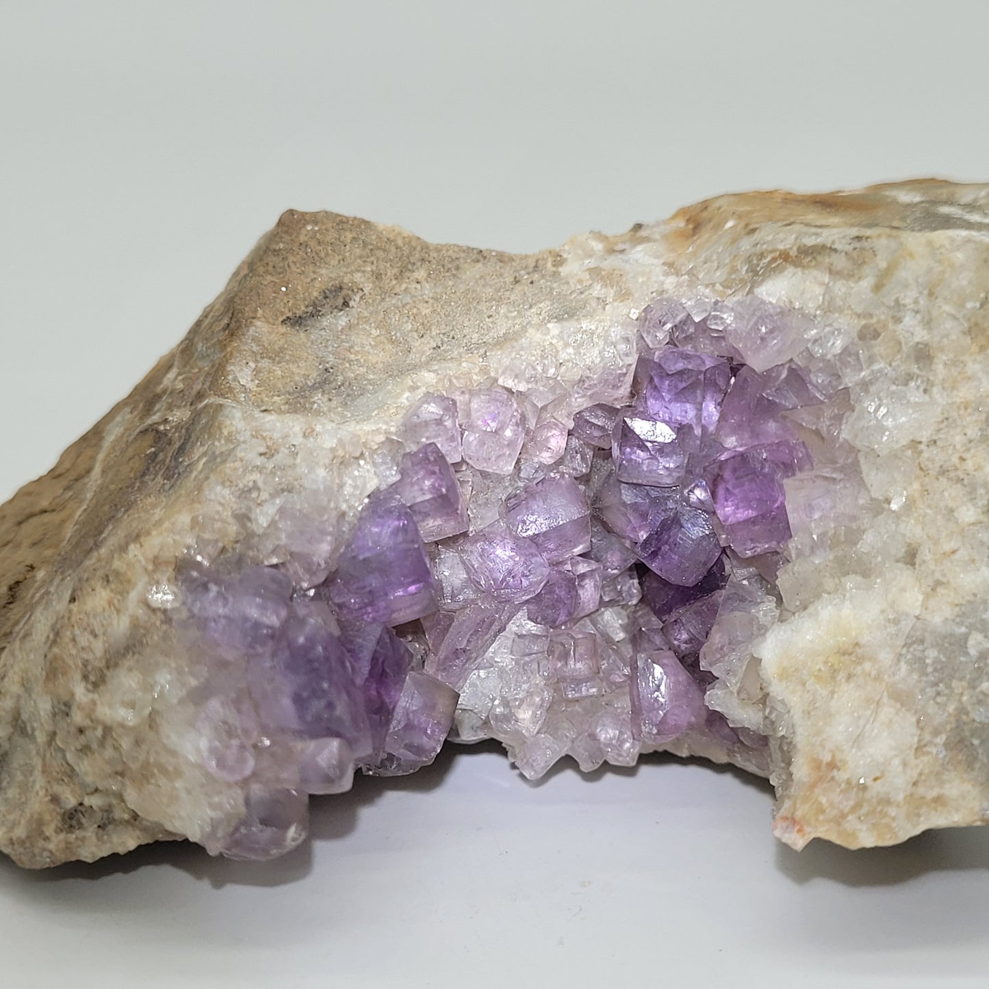 A rough geode from Jaimina Mine, Asturias, Spain (The Crystalary, sku 810) is partially split open to showcase glossy clusters of light purple amethyst crystals inside against a neutral background.