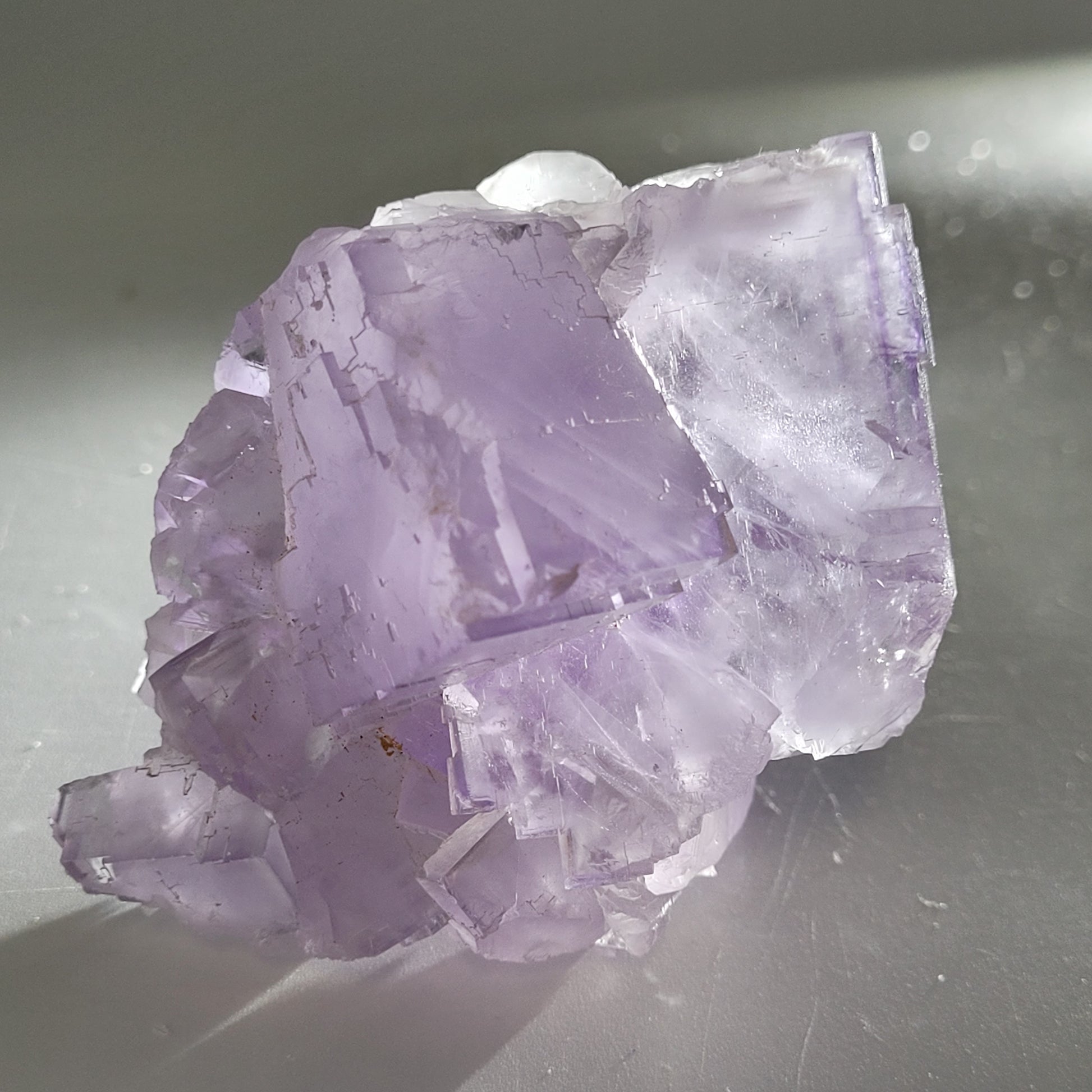 A light purple, translucent fluorite crystal cluster from Berbes (The Crystalary, SKU 811) is showcased on a smooth gray surface. Soft lighting accentuates its facets and edges, highlighting the textured surface for depth and shimmer.