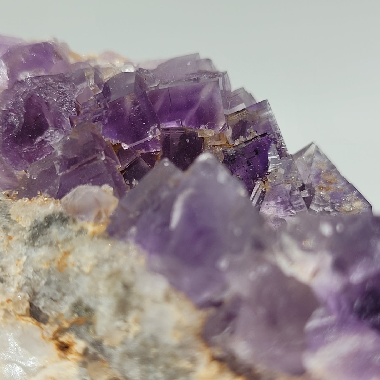 A close-up of The Crystalary product, Fluorite from Berbes, Ribadesella (sku 812), shows purple amethyst crystals with sharp edges on a rough beige rock. The renowned Ribadesella fluorite adds depth and translucency, catching light to reveal varying purple shades against an off-white background.