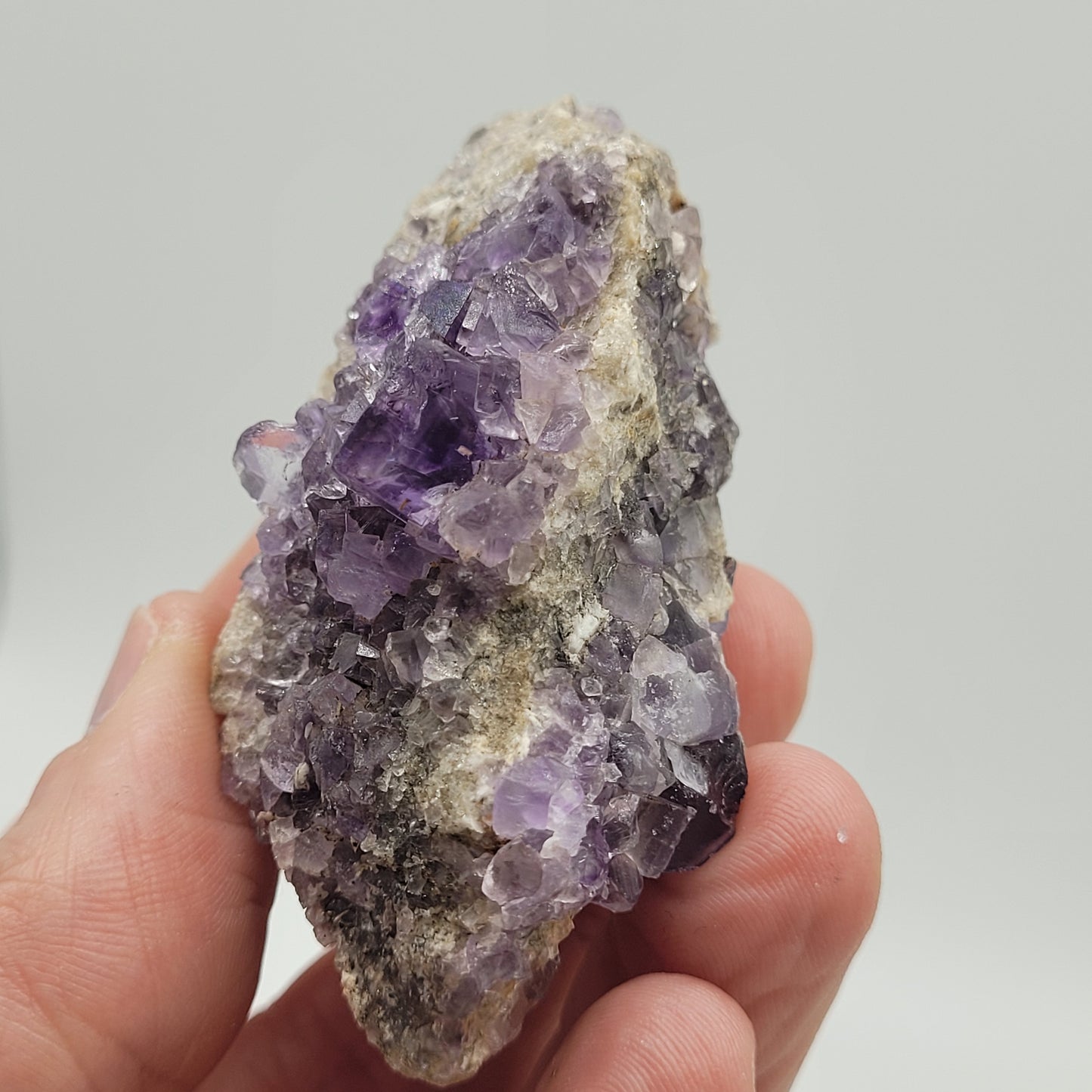 A hand displays a rough Fluorite crystal cluster from Berbes, Ribadesella (sku 813) by The Crystalary, highlighting its jagged edges and varied shades. Set against a plain background, this gem's texture and color variations vividly stand out.