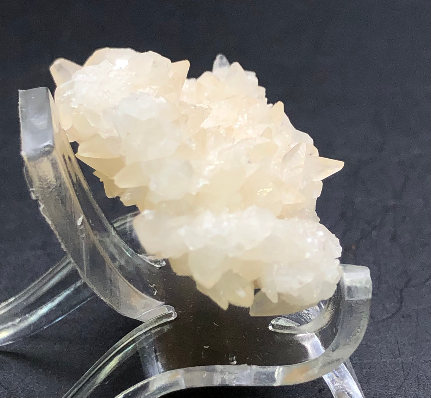 Close-up of a cream-colored calcite crystal from The Crystalary's Southwest Mine collection, displayed on a transparent stand against a dark background, showcasing sharp formations and sparkling texture reminiscent of Bisbee Arizona's peach calcite beauty.