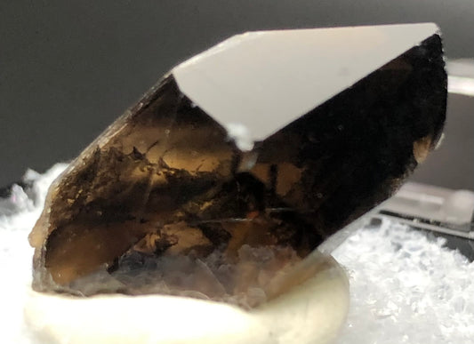 A close-up of The Crystalary's AUCTION- Smoky Quartz from Mount Malosa, Zomba, Southern Region, Malawi, showcases its glossy surface and sharply angular edges that reveal a translucent quality. The crystal rests on a white substance against a blurred and neutral background, enhancing its natural beauty.