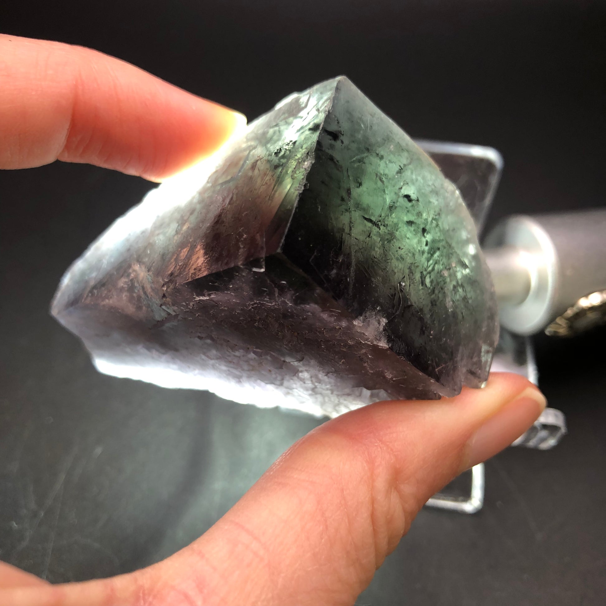 Close-up of a hand holding a large, transparent mineral sample from The Crystalary's AUCTION-Fluorite-Purple Rain Pocket, sourced from the Lady Annabella Mine in Eastgate, Weardale, Co. Durham, England. Set against a dark background, the mineral's rough and angular shape is illuminated from behind, highlighting its natural texture and clarity.