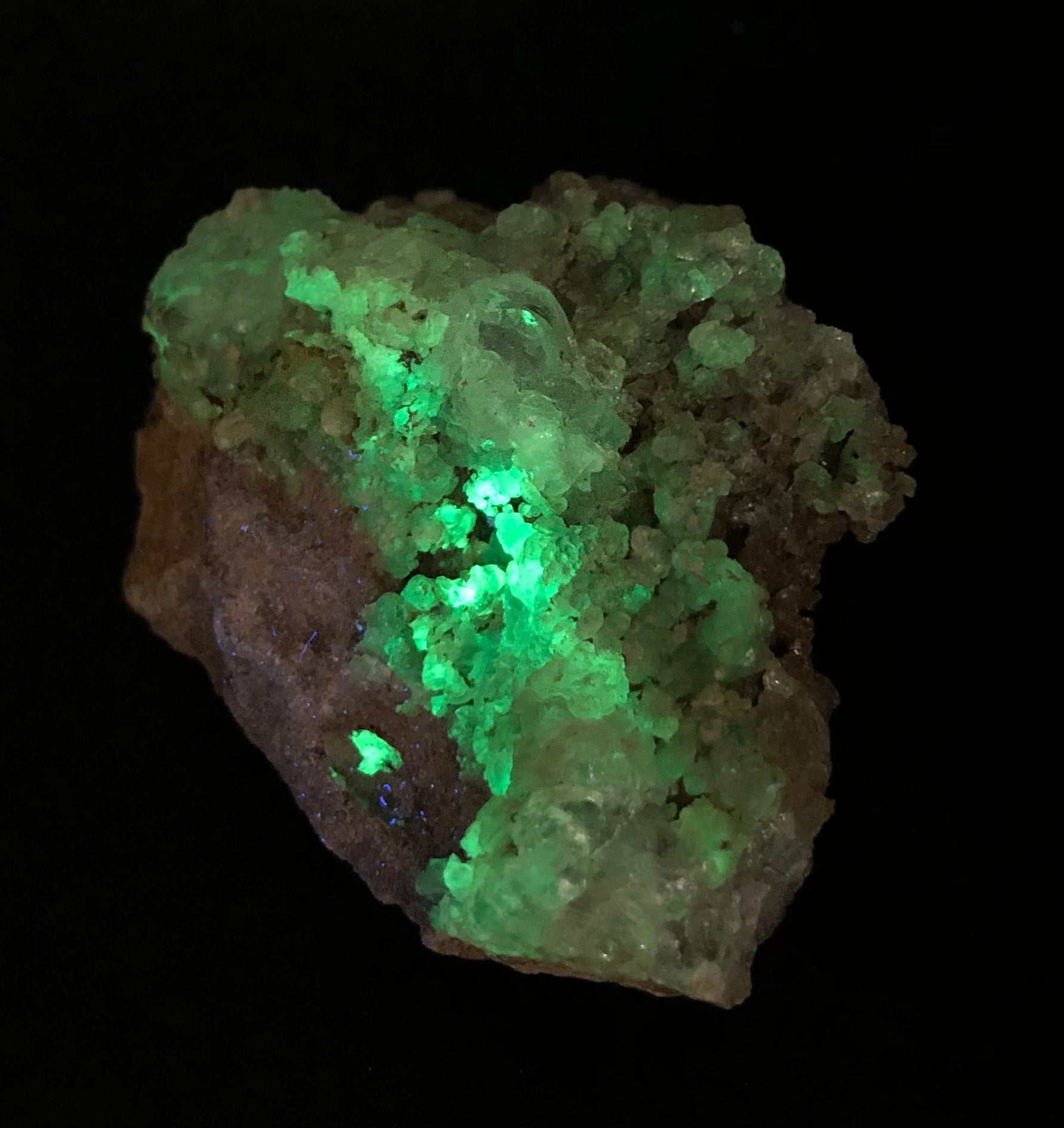 An ethereal green and blue fluorescent glow emerges from The Crystalary’s Hyalite specimen from La Ventilla, Villa de Reyes Municipality, San Luis Potosí, Mexico, accentuating its crystalline texture and uneven surface against a dark background.