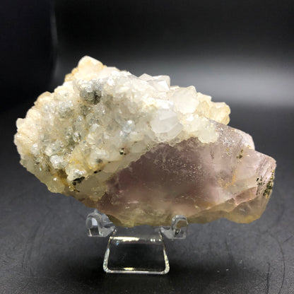 A Fluorite crystal cluster from an unknown Chinese locality, offered by The Crystalary, sits on a small stand against a dark background, showcasing its rough and intricate structure with clear and light purple hues.