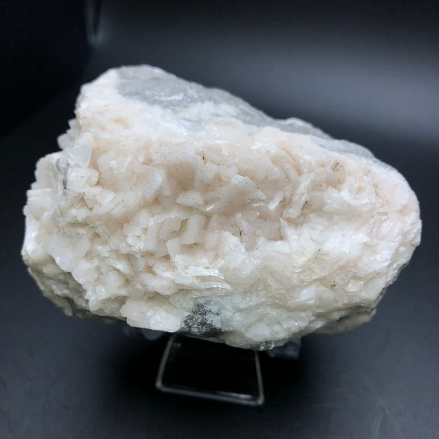 Displayed on a small stand is the AUCTION- Dolomite, Calcite mineral from Thunder Bay, Ontario, Canada. This piece by The Crystalary features a white calcite crystal cluster with a rough, jagged surface accompanied by clear dolomite crystals. The simple dark gray background highlights the texture and color of these stunning minerals.