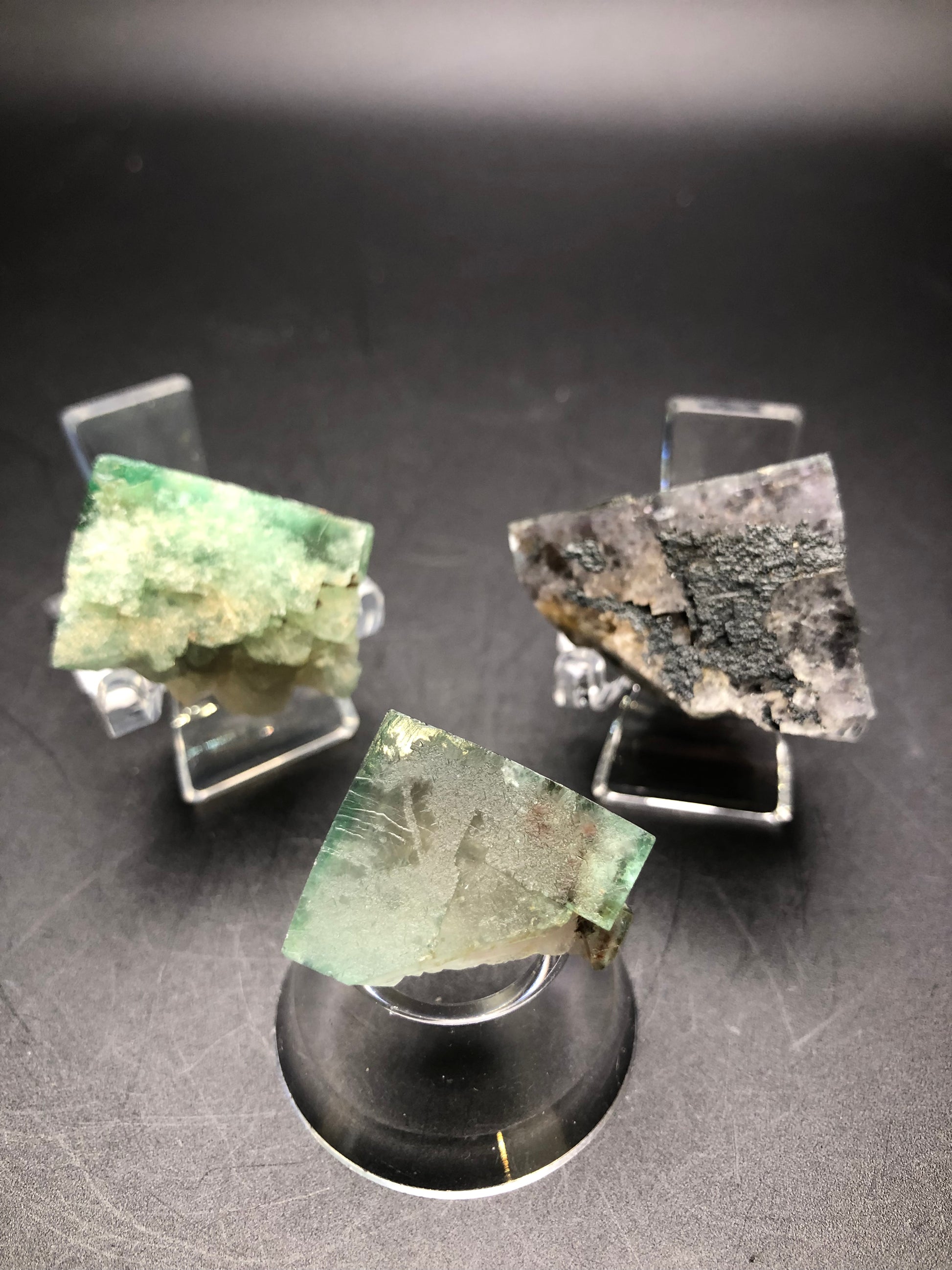 Three Fluorite mineral specimens sourced from the Lady Anabella Mine, part of The Crystalary collection, are showcased on clear stands. One specimen changes color in daylight, transitioning from green to gray, while the other two feature a striking green and dark gray in partial cube formations. These pieces are arranged in a triangular display on a dark surface.