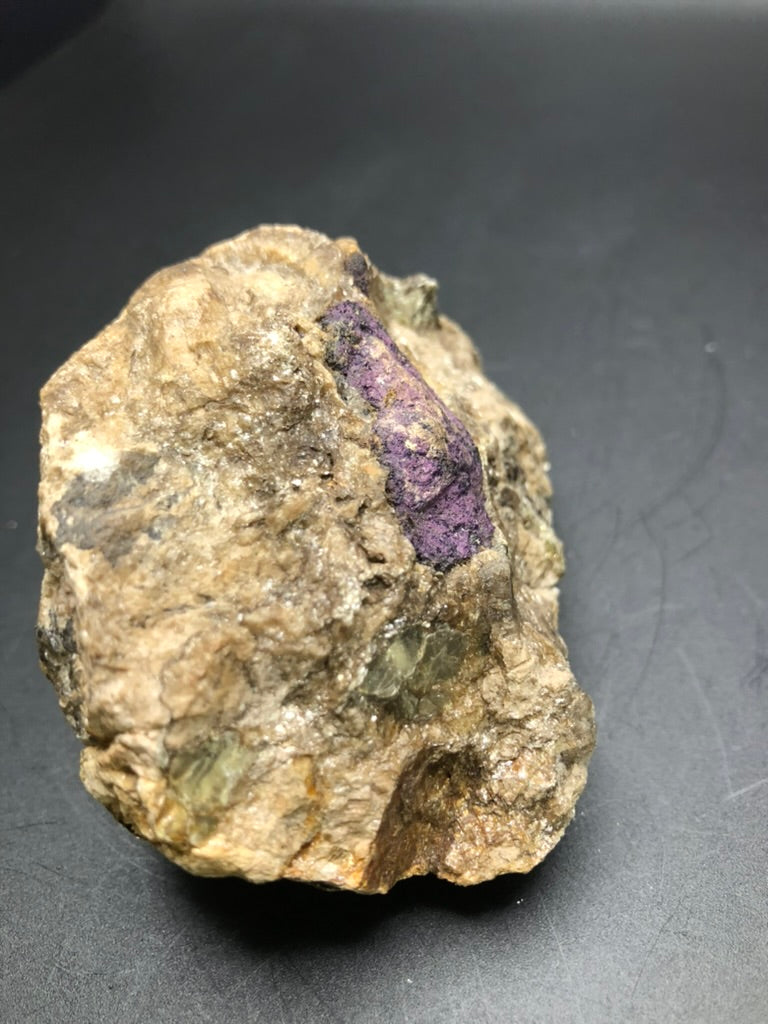 A rugged brown rock from The Crystalary, featuring a striking patch of vibrant purple Wilsonite from Canada on its surface against a dark background, makes for an intriguing mineral specimen.