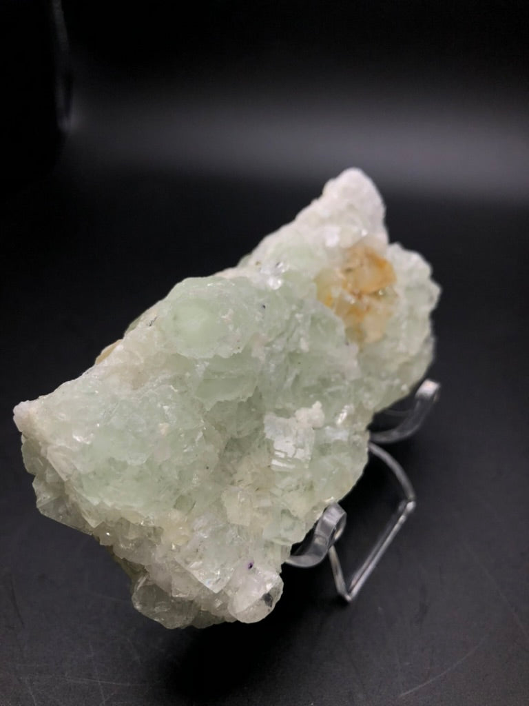 The Crystalary presents the AUCTION- Fluorite specimen from the Xianghualing Mine in Linwu Co., Chenzhou, Hunan, China. This large piece exhibits a translucent green mineral with rough, jagged edges and is elegantly displayed on a clear acrylic stand against a dark background. The crystalline texture features subtle hints of pale orange, and upon closer inspection, you can discern embedded fluorite cubes within its surface.