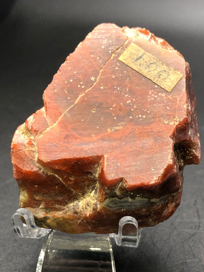 A polished reddish-brown agate specimen from The Crystalary's AUCTION collection is elegantly displayed on a clear stand against a dark background. Resembling an exquisite red agate slice, it features a small rectangular label with faded writing on its surface.