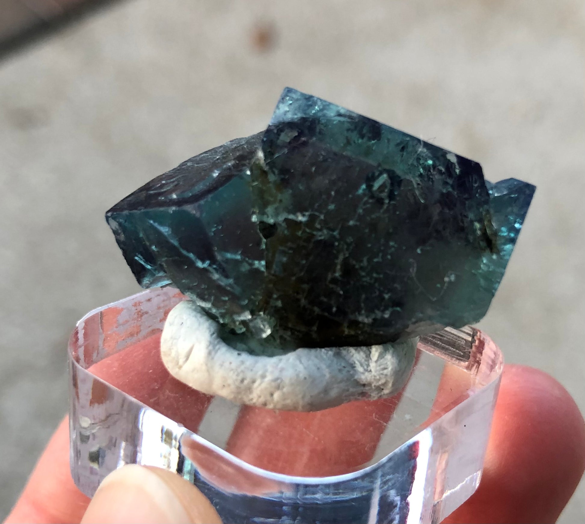 A close-up image featuring "AUCTION- Fluorite" from The Crystalary highlights a dark green crystal sourced from the Diana Maria Mine in Frosterley, Weardale, County Durham, England. Mounted on a clear acrylic base, the rough, geometric shape of the fluorite is showcased against a softly blurred neutral background as it is held between two fingers.