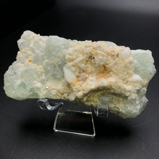 Displayed on a clear stand, the mineral specimen from The Crystalary, titled "AUCTION- Fluorite- Xianghualing Mine, Linwu Co., Chenzhou, Hunan, China," showcases its pale green and white hues with captivating fluorite cubes. Its rough texture and crystalline formations offer a striking visual contrast against a dark background, reflecting its origins in the Xianghualing Mine.