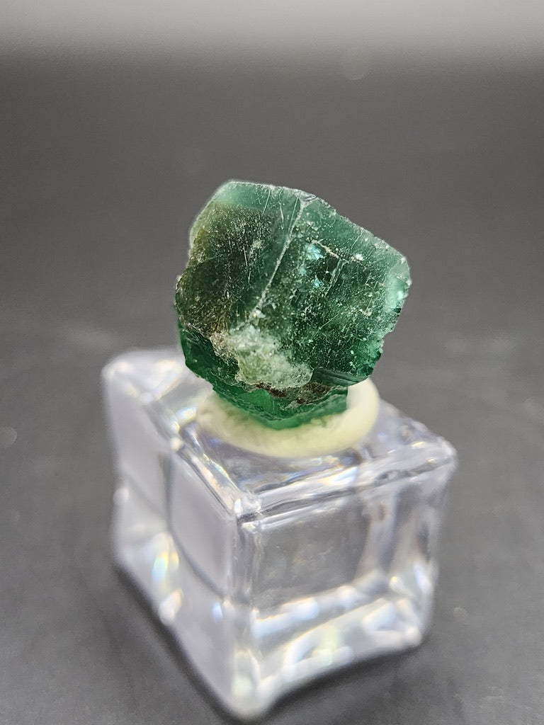 Positioned on a transparent square stand against a dark background, the rough green fluorite gemstone from the Greedy Hog Pocket showcases its natural texture and color variations. This captivating piece from The Crystalary originates from Diana Maria Mine in Weardale, Co Durham, UK, and mesmerizes with its unique sunlight fluorescence.