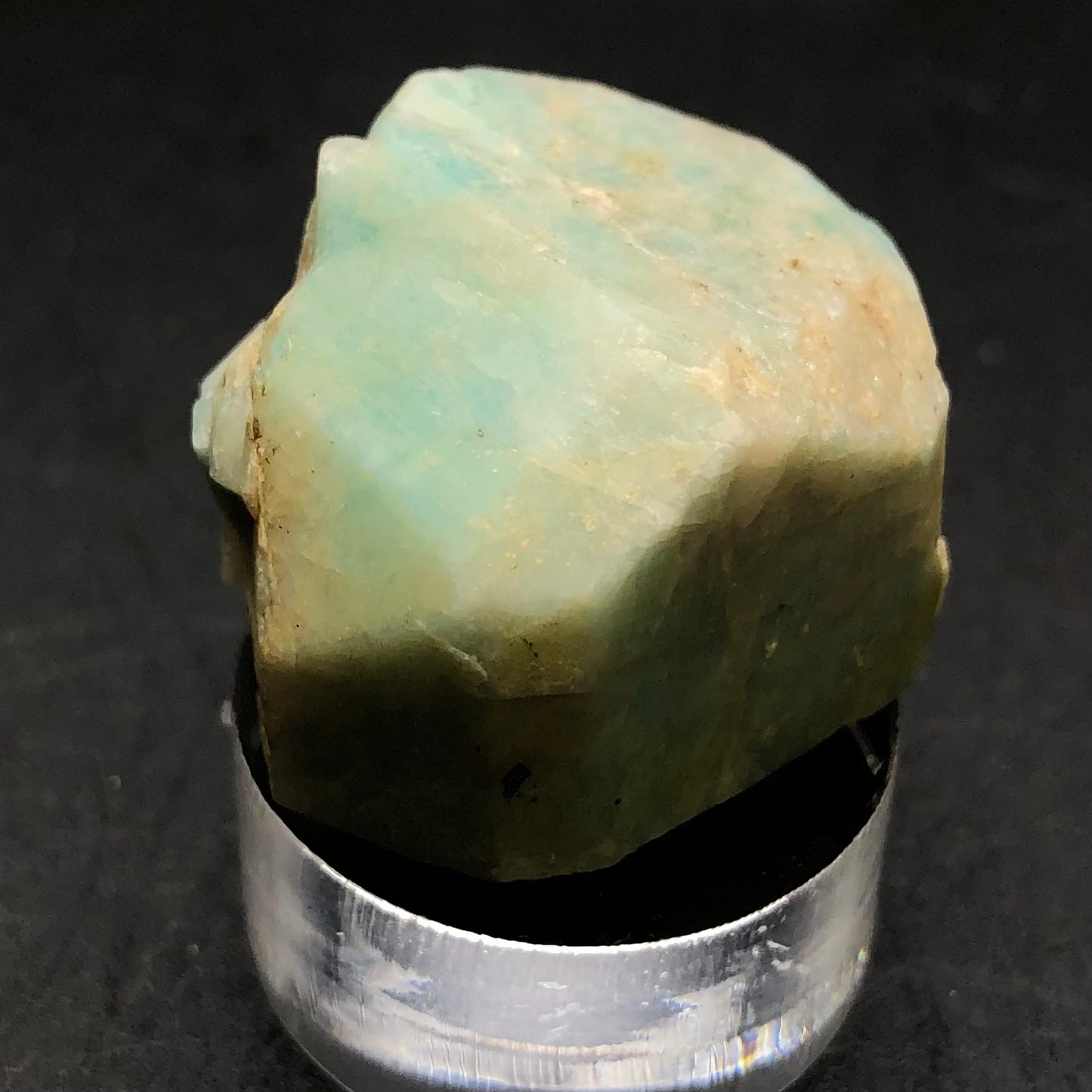 The Crystalary introduces their Amazonite specimen sourced from Lake George, Park County, Colorado, USA. This rough stone displays a pale green hue with a vitreous luster and uneven surfaces due to its triclinic crystal structure. It is elegantly presented on a round silver base against a stark black background.