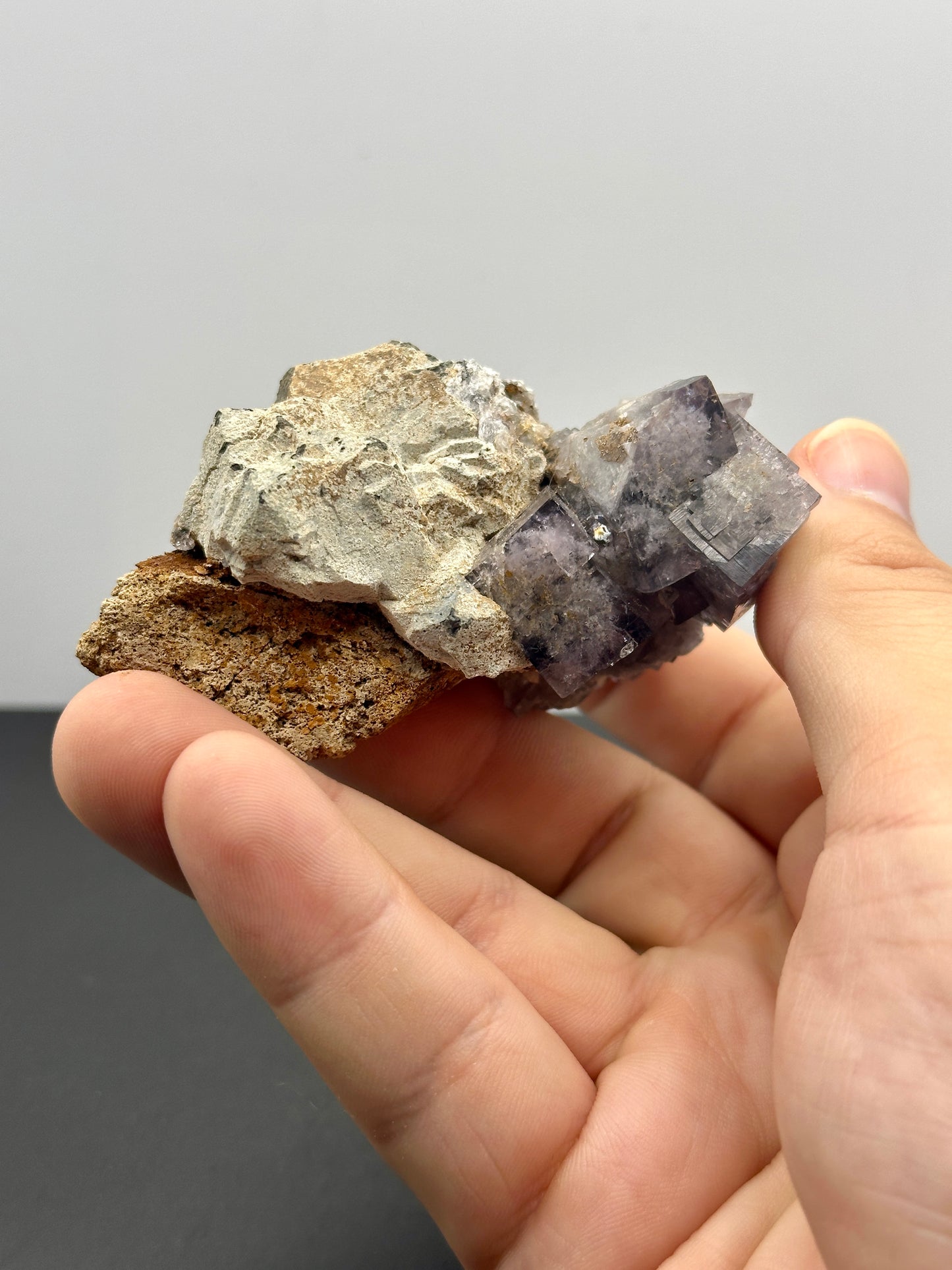 A hand showcases a mineral specimen from The Crystalary's collection, specifically the Fluorite from the Hailstorm Pocket at the Lady Annabella Mine, featuring fluorescent purple crystals accented by rough, earthy materials. The backdrop is a neutral gray surface.