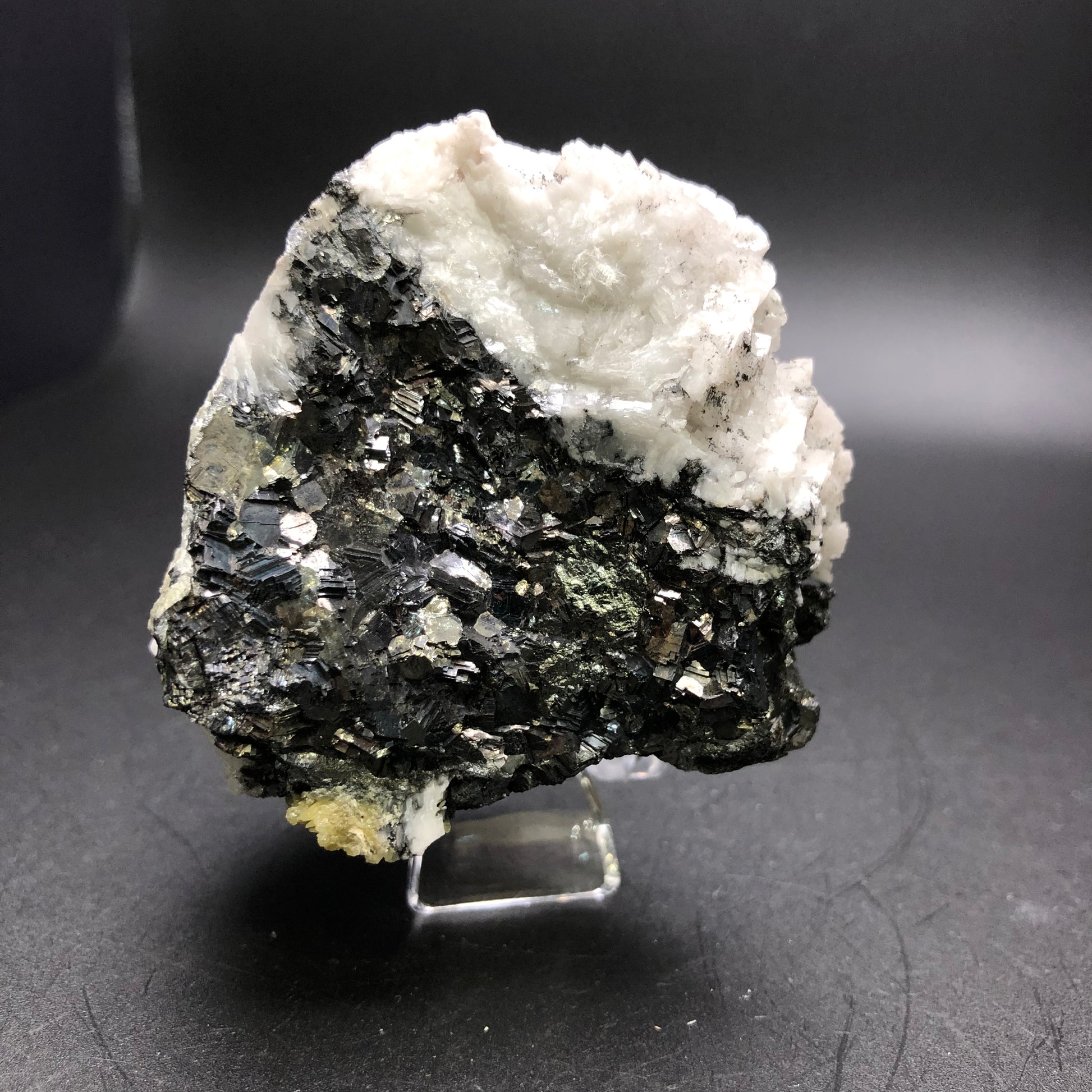 A striking piece from The Crystalary, the Mimetite and Dolomite on Galena from the Tsumeb Mine in Namibia showcases shiny metallic surfaces set against a dark backdrop. Adorned with white, textured dolomite formations on its upper section, this highlights a contrast to the lustrous black base and is elegantly presented on a small, clear stand.