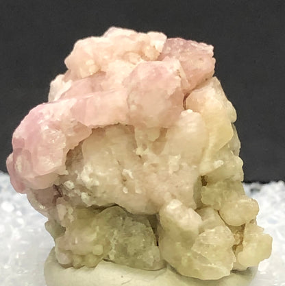 The Crystalary's AUCTION - Garnet, Grossular from Morelos, Mexico features a close-up image of light pink garnet crystals clustered on a rocky base against a dark background, highlighting their intriguingly rough and uneven texture.