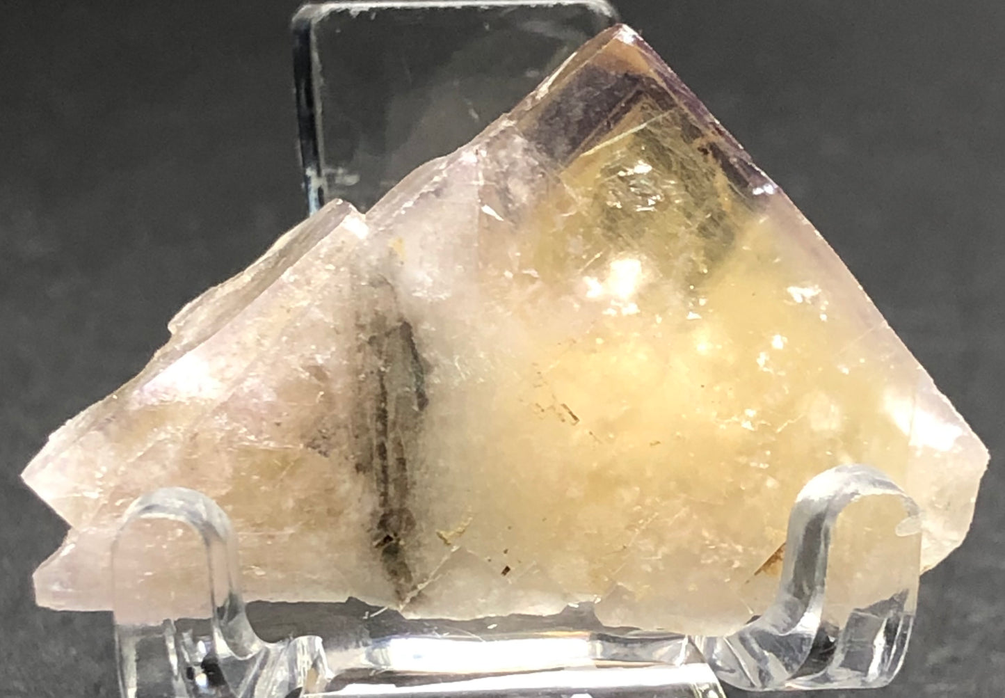 A close-up of The Crystalary's AUCTION- Fluorite from Cromwell Pocket, Greenlaws Mine highlights its translucent light yellow color with white and purple hues. Its vitreous partial cube form showcases golden interior tones and stunning purple exterior zoning against a dark background.