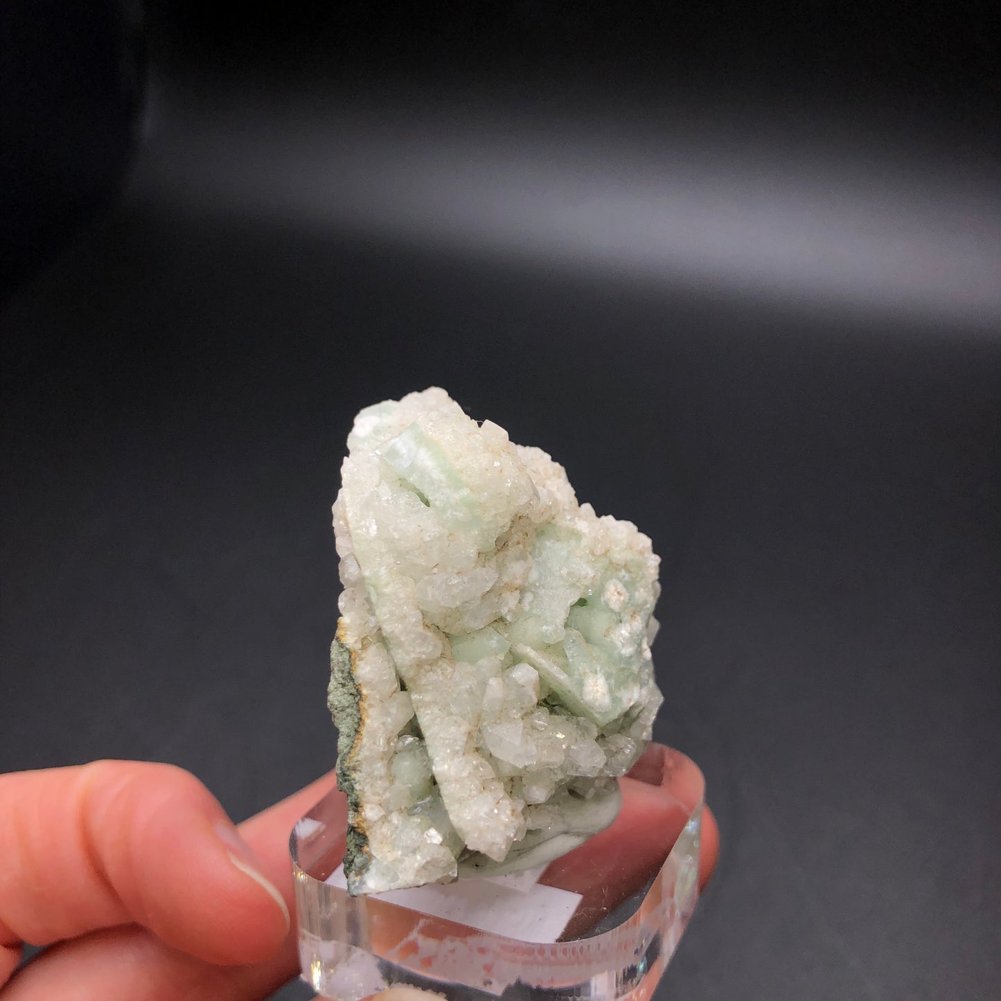 A hand holds a dazzling cluster of The Crystalary's Apophyllite on Prehnite from the D&G Quarry in Woodbury, Litchfield County, Connecticut. It sits elegantly on a clear stand against a dark background, showcasing minty green and white hues with a rough, jagged texture.