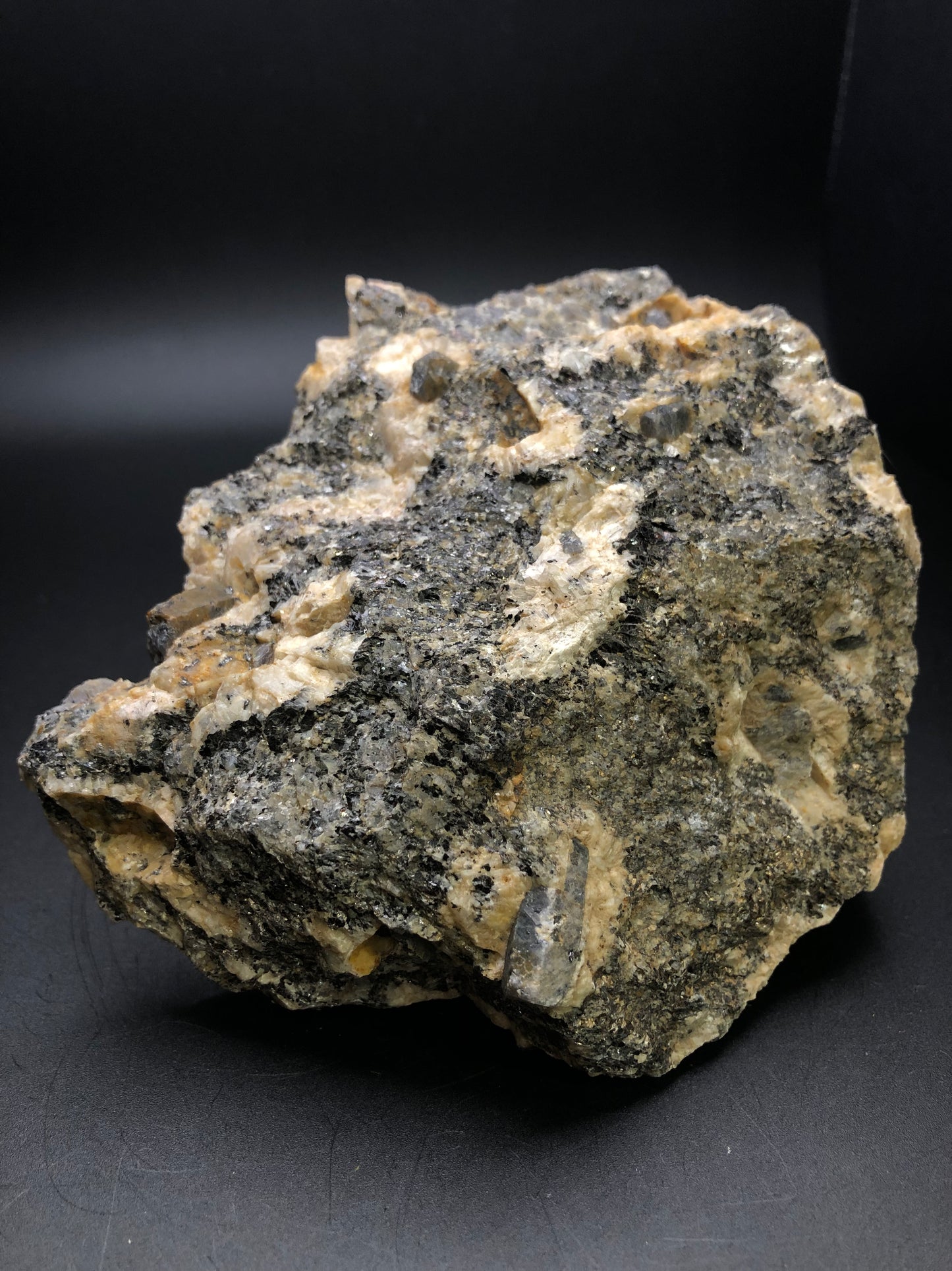 The Crystalary's AUCTION- Corundum from the Elk Creek Corundum Deposit in Gallatin County, Montana features a rough, irregularly shaped rock with a textured surface of gray and tan hues against a black backdrop, adorned with small corundum crystals scattered throughout.