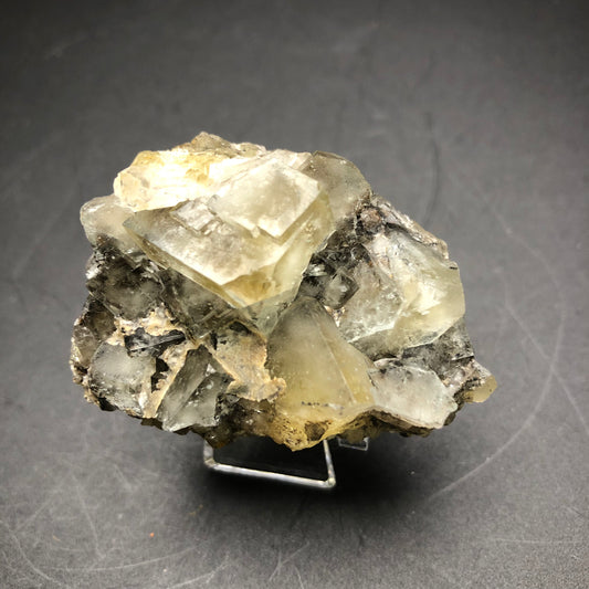 A close-up of "AUCTION- Fluorite" by The Crystalary, sourced from The Bull Vein at the Lady Annabella Mine in Eastgate, Weardale, Co. Durham, England, reveals its stunning gray and beige crystalline formations. Set on a small metal stand against a dark background, this exquisite specimen showcases unique daylight color-changing properties.