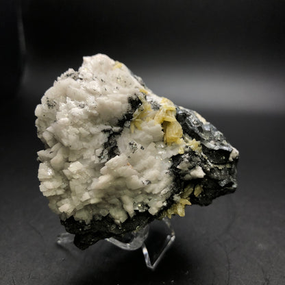 The Crystalary product, "AUCTION - Mimetite, Dolomite on Galena" from the Tsumeb Mine in Namibia, showcases a detailed view of a mineral specimen featuring a cluster of white, blocky crystals accented with black and yellowish tones. The display is set against a dark backdrop.