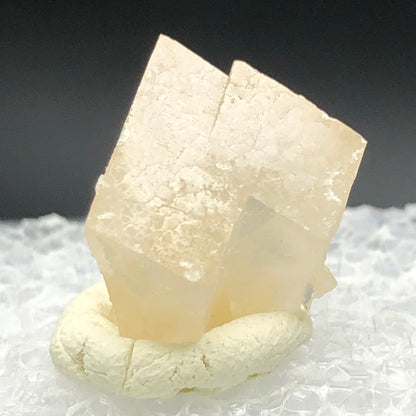 A translucent, light honey calcite crystal with a cubic structure from Otero County rests on a textured white surface, set against a soft, dark background that highlights its intricate details and natural patterns. Product: AUCTION- Calcite- Otero County, Colorado, USA by The Crystalary.