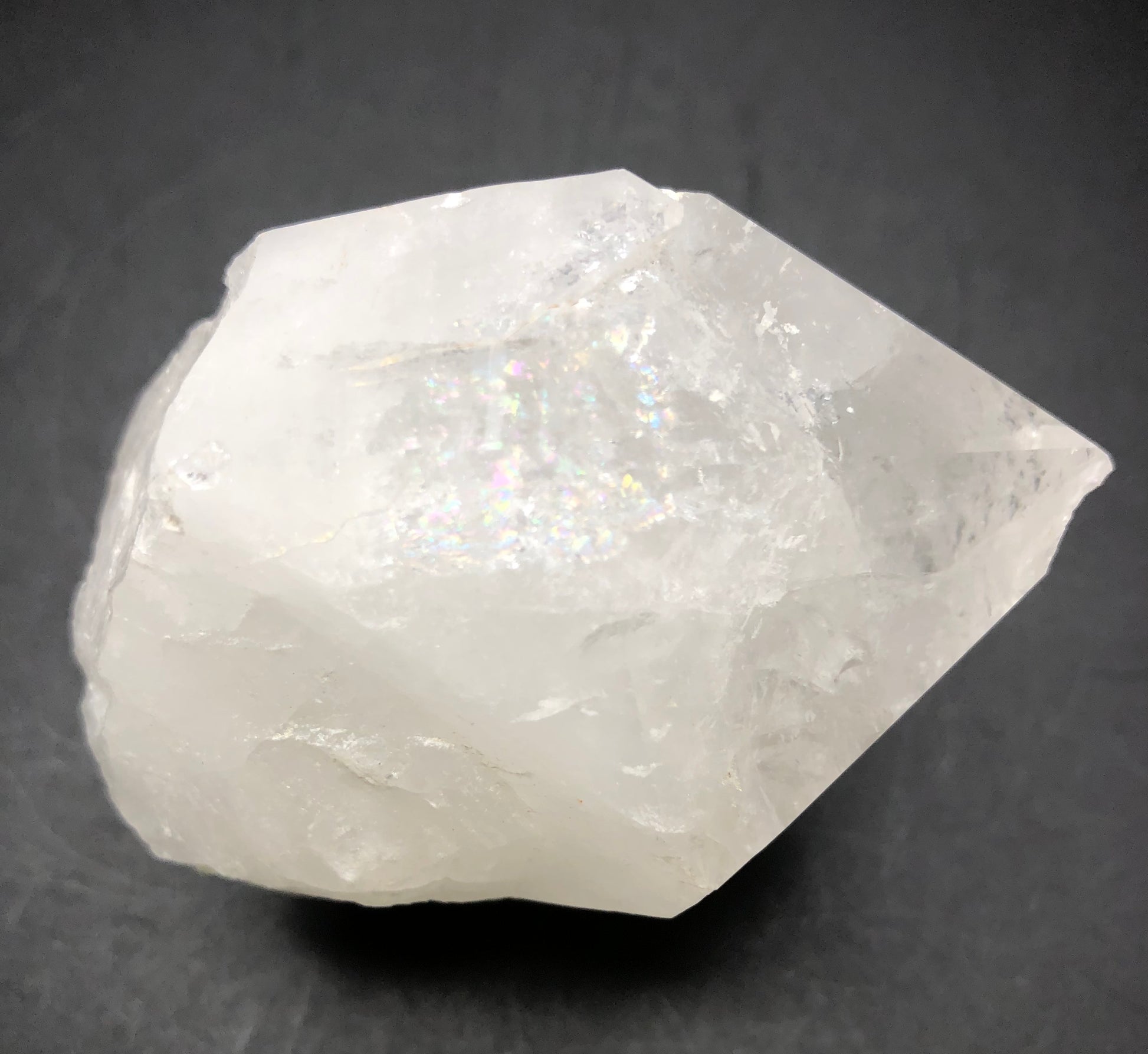 A large, clear quartz point from The Crystalary's AUCTION - Quartz - unk locality collection lies against a dark background. Its rough texture and noticeable facets highlight its transparency and reflective, light-catching areas.