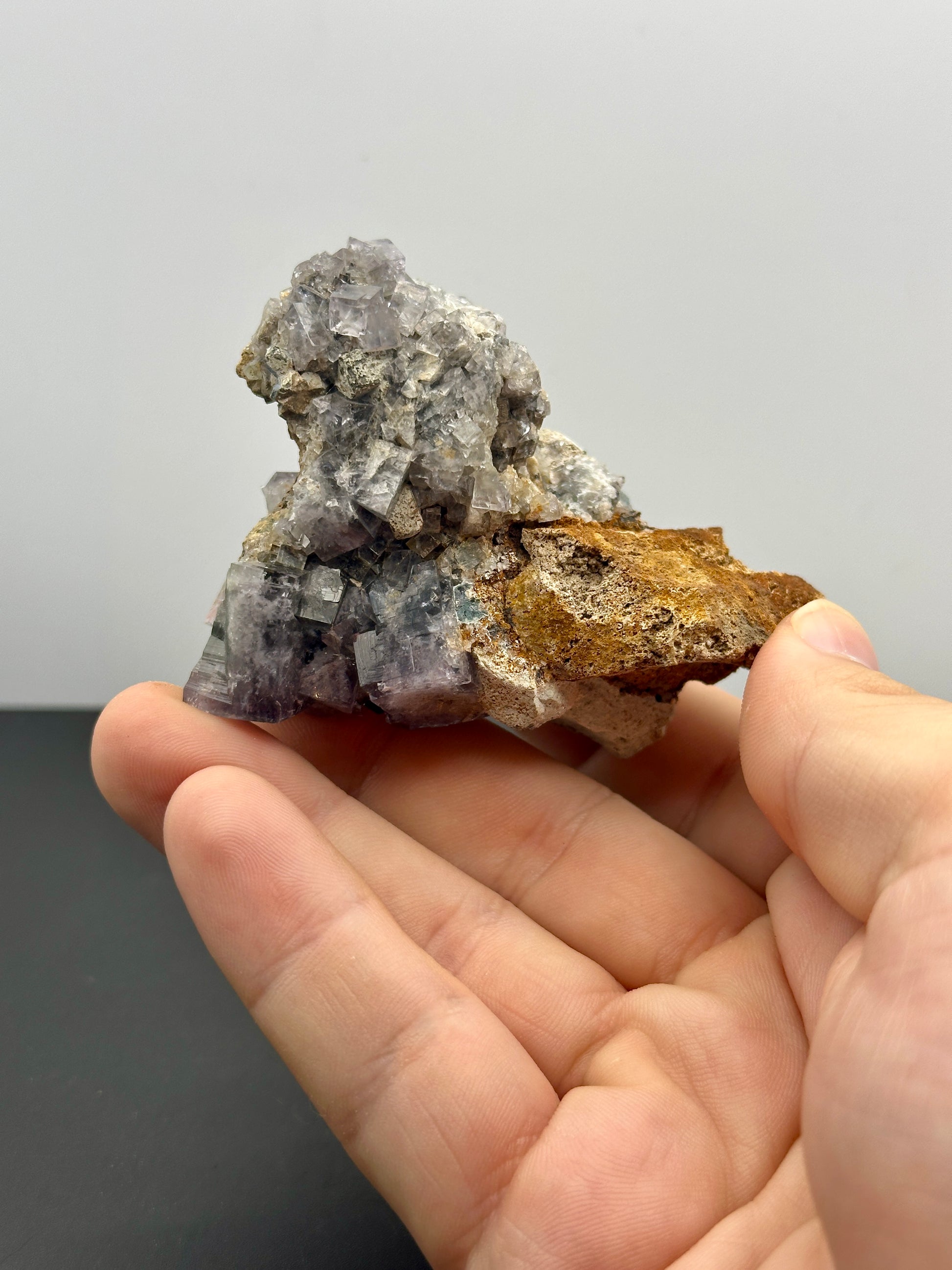 A hand displays a mineral specimen from The Crystalary, featuring the Fluorite- Hailstorm Pocket (HS001) from the Lady Annabella Mine in Weardale, Co Durham, UK. The cluster of translucent cubic crystals rests on a rusty brown rock base and illuminates with a striking fluorescent purple hue under certain light conditions, all set against a plain background.