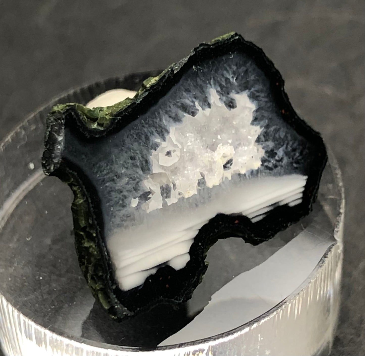 Close-up of "AUCTION- Tabasco geode- Tabasco, Mexico" by The Crystalary, featuring a polished, cross-sectioned geode with dark outer layers and a bright, crystalline center. It is presented on a circular, transparent platform set against a gray background.