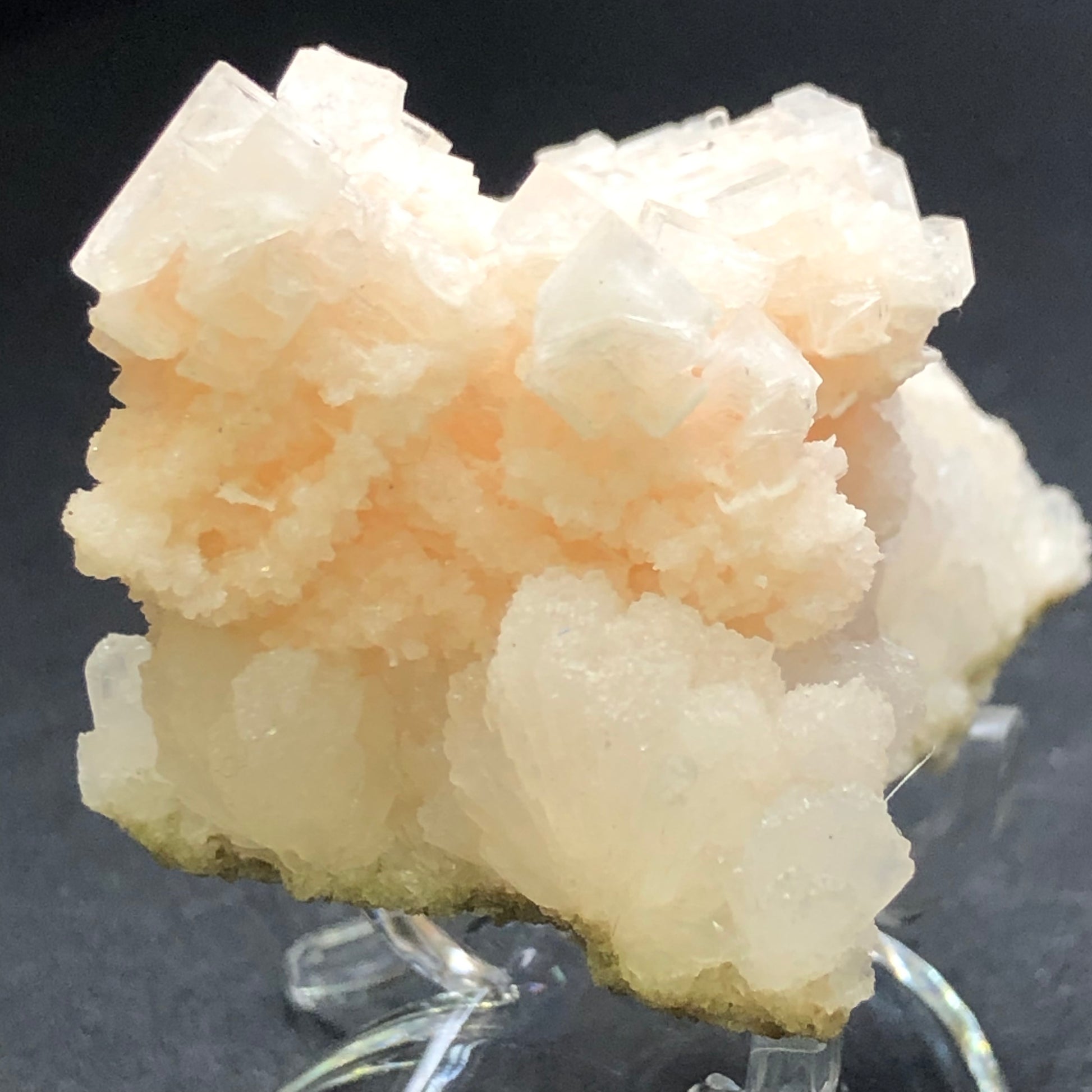 A close-up of a light pink stilbite crystal formation showcases its intricate, layered textures against a dark background. The delicate structure and shimmering surface evoke the geometric beauty synonymous with the Halite crystals offered by The Crystalary in their AUCTION- Halite- unk locality collection.