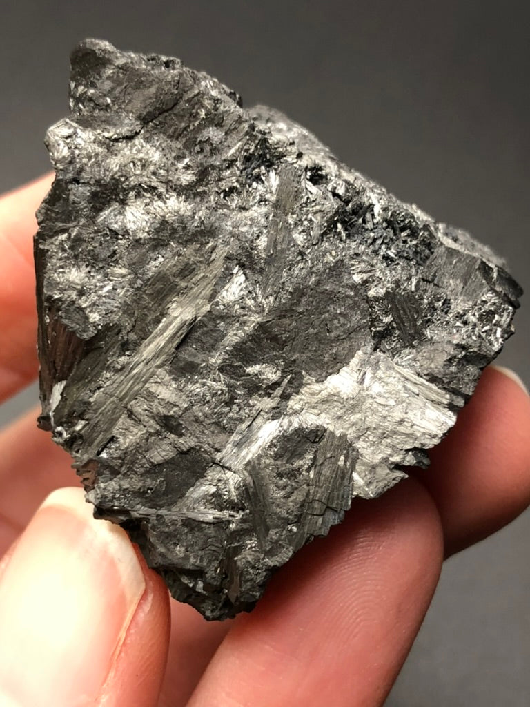 A close-up view captures a hand holding a rough, metallic mineral with a shiny, dark gray surface. Its radiating crystals create a layered and jagged texture that reflects light beautifully against a plain, dark background. This could be the "AUCTION - Pyrolusite" from Elgersburg, Ilm District, Thuringia, Germany by The Crystalary.