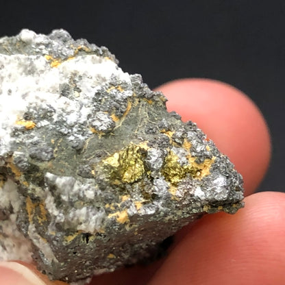 A close-up of a hand holding The Crystalary's "AUCTION- Pyrite, hematite- unk locality" mineral specimen reveals visible gold veins against a dark background, featuring gray stone and golden metallic spots, suggesting an intriguing pyrite composition.