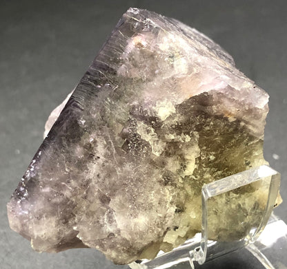 A close-up image of the translucent fluorite mineral from The Crystalary, sourced from Cromwell Pocket, Greenlaws Mine, UK, shows a mix of purple and gray hues. Displayed on a clear stand against a dark background, its uneven surfaces reflect light beautifully and hint at unique daylight color-changing properties.