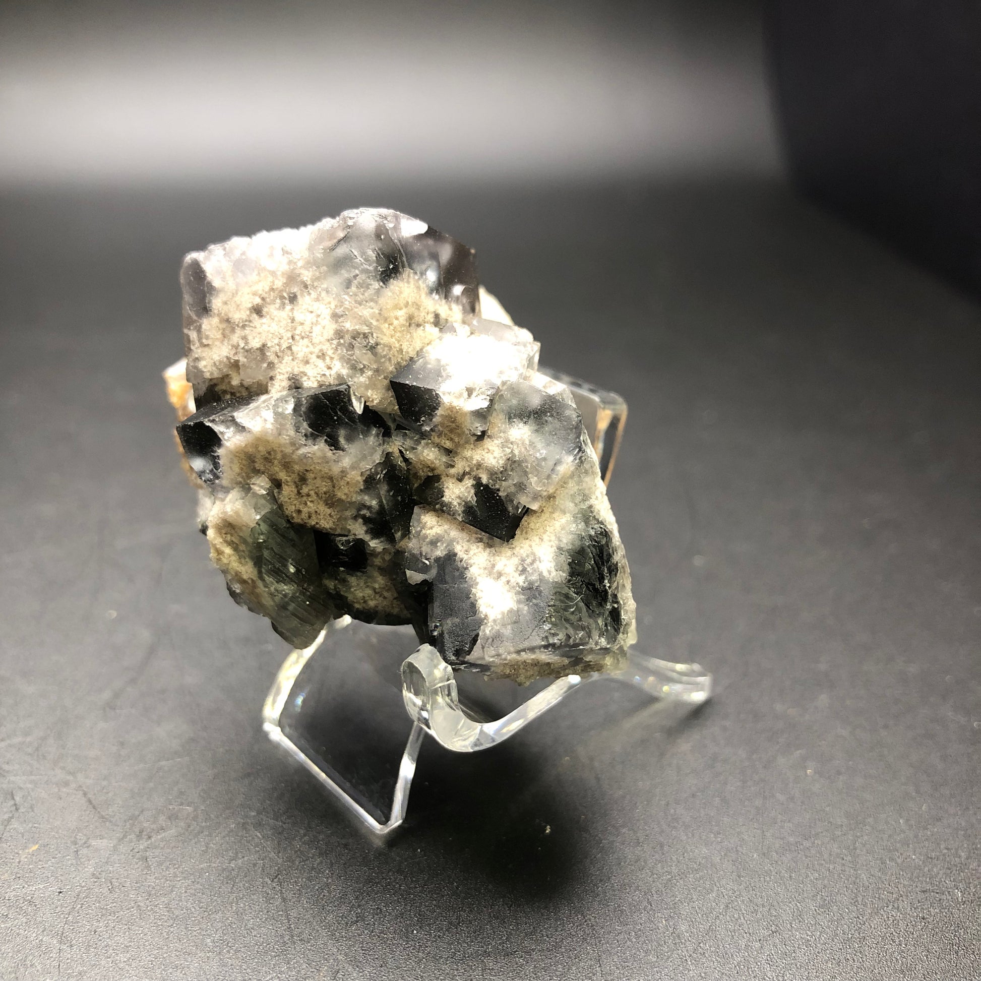 A cluster of black and white crystal formations from "AUCTION- Fluorite- Milky Way Pocket, Diana Marie Mine, Frosterly, Weardale, Co. Durham, England" by The Crystalary is displayed on a clear stand against a dark background. The smoky gray-blue fluorite crystals exhibit a rough, jagged texture and shine glossily under the light.