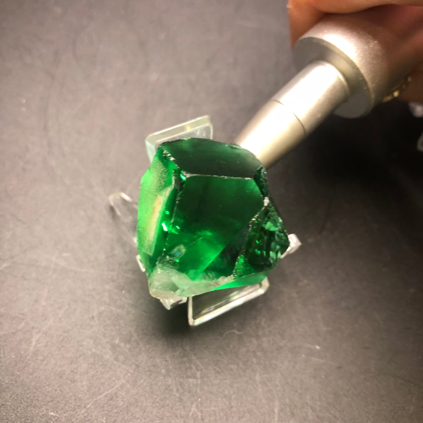 A vibrant deep green fluorite gemstone, sourced from the Diana Maria Mine in Frosterly Weardale Co., Durham, England, is elegantly held by a silver tool and rests on a black surface. This multifaceted fluorite cube from The Crystalary shines brilliantly with its glossy finish.