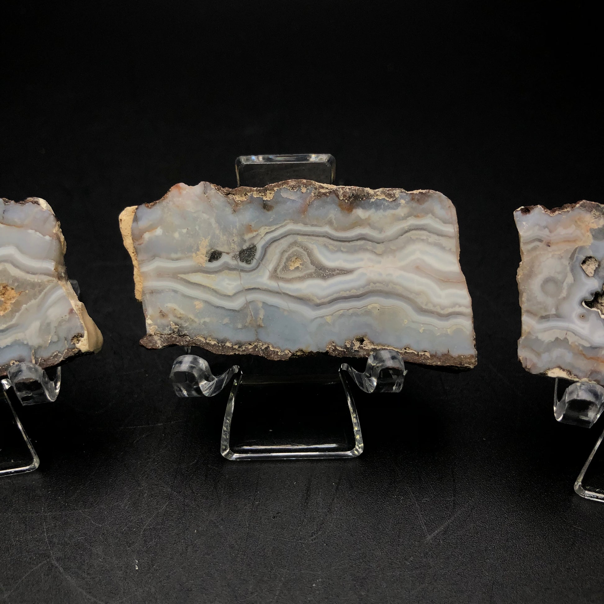 Displayed on a clear stand against a black backdrop, a slice of Arizona agate from The Crystalary's "AUCTION- Agate" collection reveals intricate bands of gray, cream, and white with its natural wavy patterns. Partially in view are two other agate slabs flanking it.