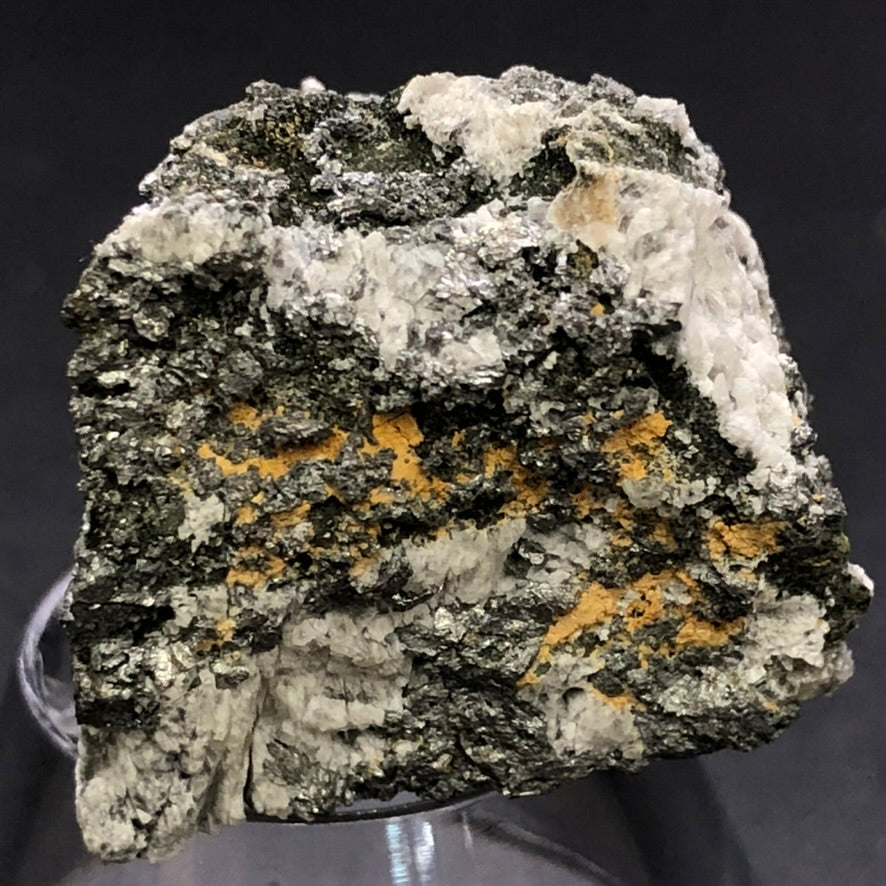 A close-up of a mineral specimen from The Crystalary's "AUCTION- Pyrite, hematite- unk locality" showcases its rough textures and dark, metallic surface reminiscent of hematite. White and orange patches adorn the mineral, displayed on a clear stand against a black background.