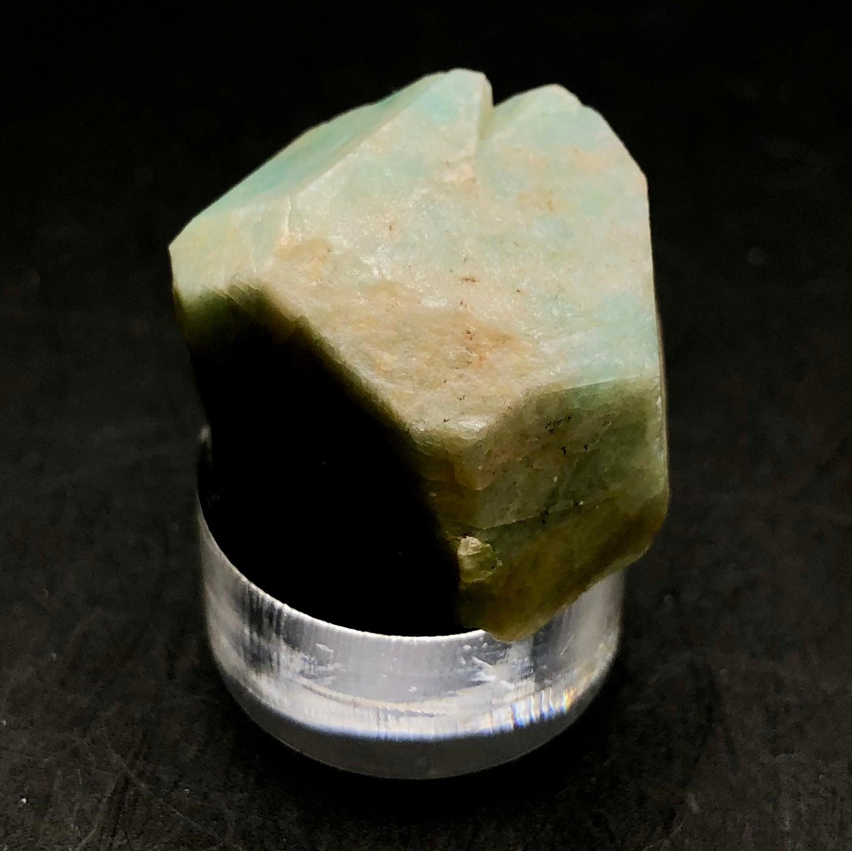 A beautifully irregular amazonite mineral from the Lake George area in Park County, Colorado, presented by The Crystalary, is displayed on a transparent circular stand against a dark background. Its surface boasts natural textures with subtle hints of brown and light blue that highlight its triclinic crystal structure.