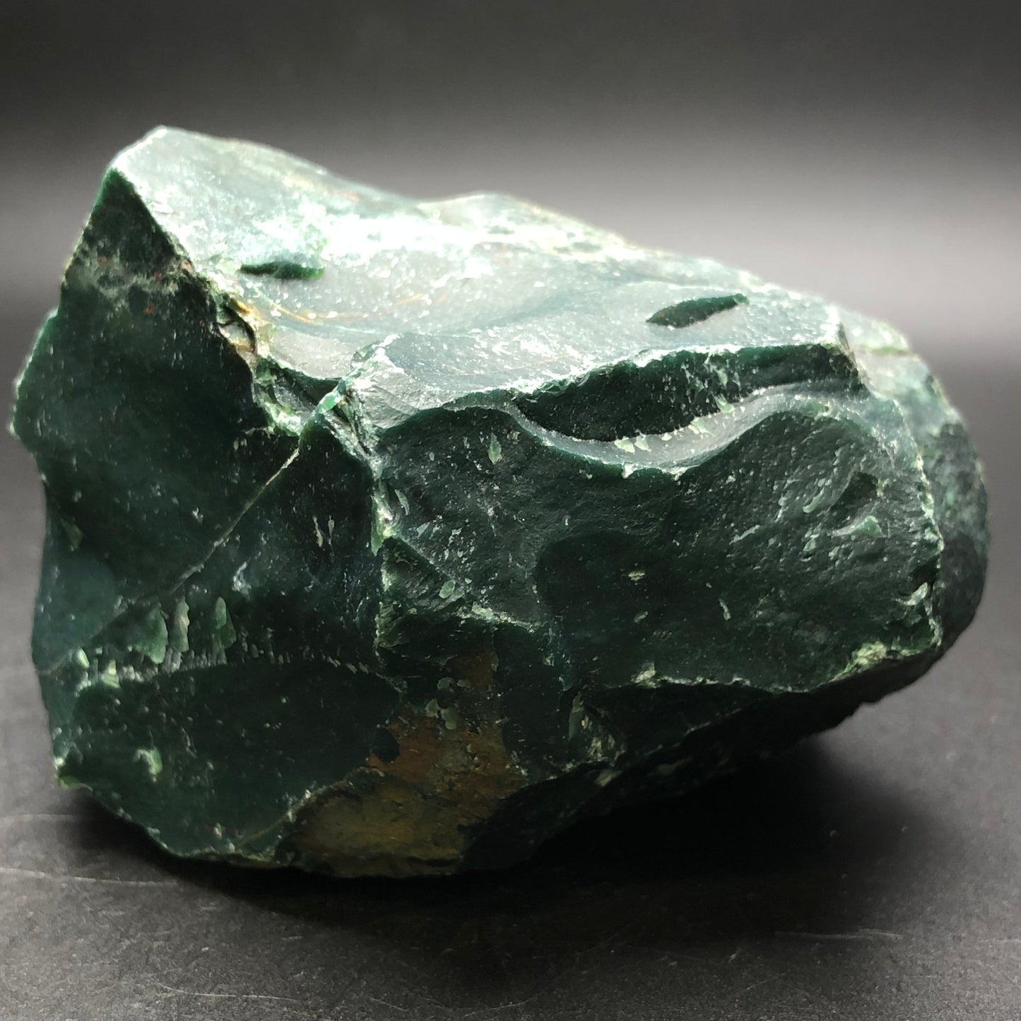 A rough, dark green bloodstone with uneven surfaces and jagged edges from The Crystalary's AUCTION- Bloodstone- unk locality, displayed on a plain black background.