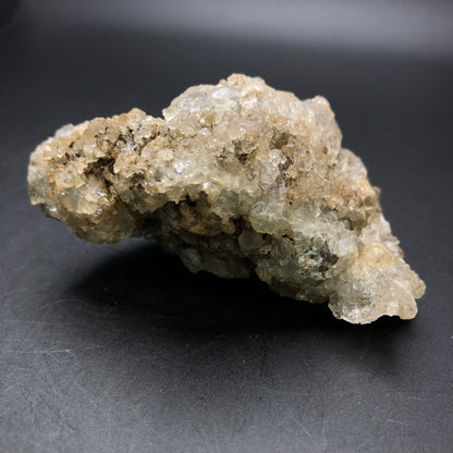 A rough, translucent mineral specimen featuring clusters of small crystals is predominantly light brown and off-white. This stalactic hyalite opal from La Ventilla, Villa de Reyes Municipality, San Luis Potosí, Mexico is highlighted against a dark background and emits a subtle green fluorescence under UV light. Available as AUCTION - Hyalite by The Crystalary.