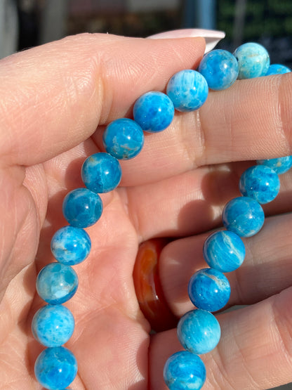 Close-up of a hand holding an 8mm Blue Apatite- Blue Friendship Bracelet by The Crystalary. The beads, measuring 8mm in diameter, are round with a marbled pattern that showcases varying shades of blue and white. The slightly blurred background enhances the focus on the vibrant bracelet.