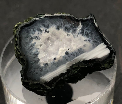 Close-up of the AUCTION- Tabasco geode from The Crystalary, showcasing its dark outer layer and dazzling druzy quartz interior. It rests on a reflective surface, accentuating its textures and colors.