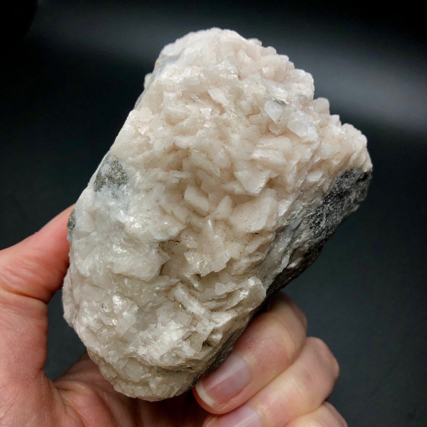 A hand displays a substantial white crystal specimen, most likely from the AUCTION- Dolomite & Calcite collection by The Crystalary, sourced from Thunder Bay, Ontario, Canada. The rock's rugged surface features small, gleaming facets that catch the light against a dark backdrop.