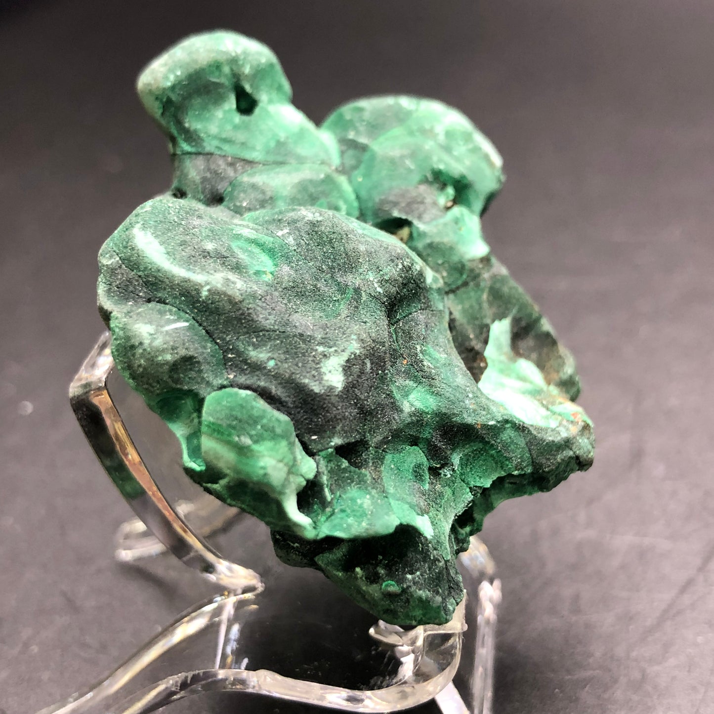 A rugged, dark green mineral specimen of raw malachite, highlighted by lighter green patches, is showcased on a clear display stand against a dark backdrop. This remarkable piece from The Crystalary was mined at the renowned Liufengshan Mine in the Guichi District of Chizhou, Anhui, China and features an irregularly textured surface.