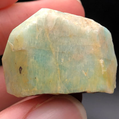 A close-up of someone's fingers holding a rough AUCTION-Amazonite gemstone from Lake George, Park County, Colorado, USA. The stone, featured by The Crystalary, showcases a mix of blue, green, and pale yellow hues with an irregular angular shape. Its slightly cloudy surface is enhanced by its natural vitreous luster.