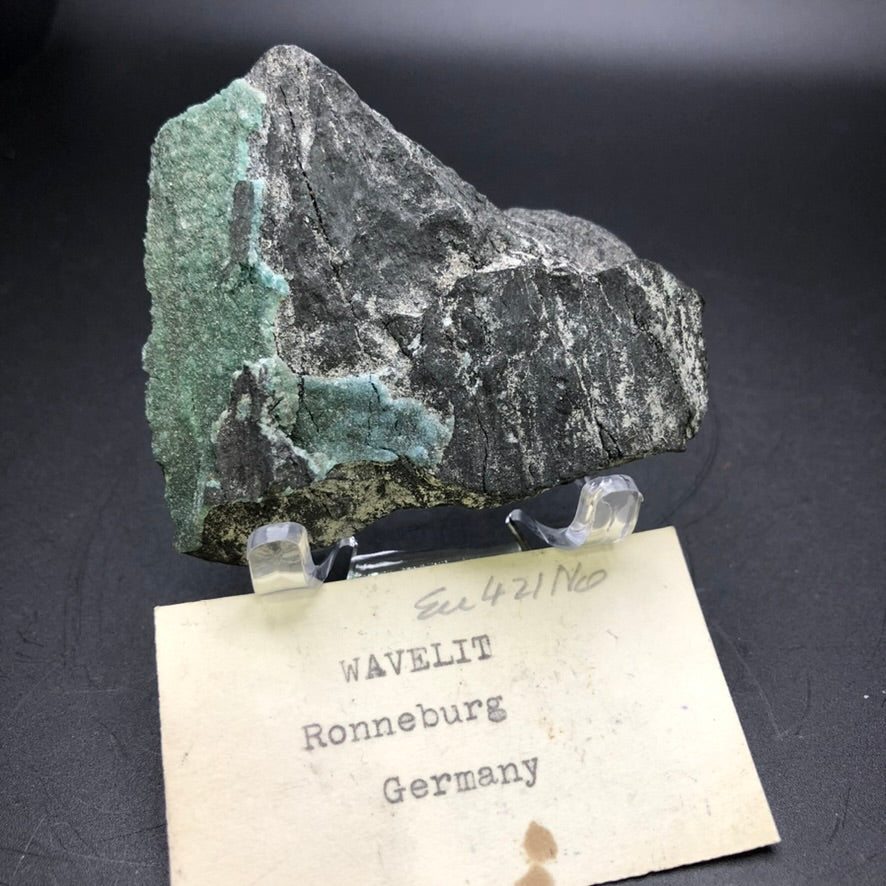 A specimen from The Crystalary's auction collection, labeled as Wavellite from Ronneburg, Gera, Thuringia, Germany, features a rough gray and green mineral surface. It is displayed on a clear stand with a handwritten note beneath it, contrasting against the plain dark background.