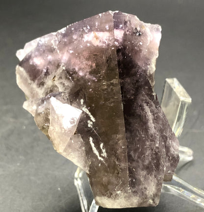 A close-up image of a large fluorite crystal from The Crystalary, featuring striations and light reflections highlighting its translucency. The AUCTION- Fluorite is showcased on a clear stand against a dark background, sourced from Cromwell Pocket, Greenlaws Mine, County Durham, UK.