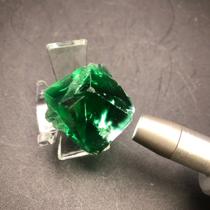 A close-up of an AUCTION- Fluorite crystal from The Crystalary, originating from the Diana Maria Mine in Frosterly Weardale Co., Durham, England, elegantly perched on a clear stand. A metallic pen-like object is pointed at the deep green crystal, highlighting its smooth and shiny cubic surfaces against a dark background.