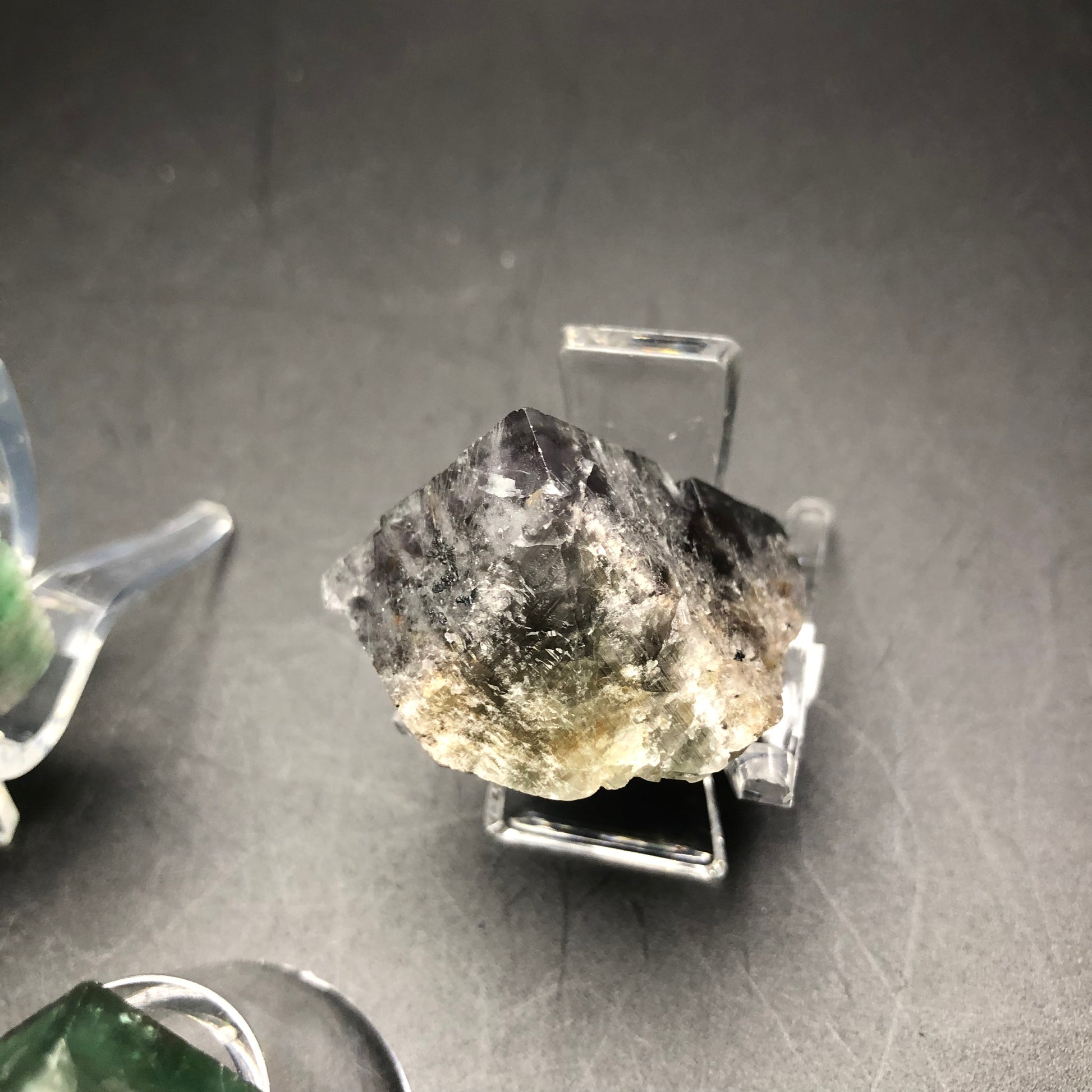 Displayed on a transparent stand, The Crystalary offers an impressive large fluorite specimen from the Lady Anabella Mine. This mineral, presented against a plain dark background, features a rough, multi-faceted surface with partial cubes in dark gray adorned with lighter patches.